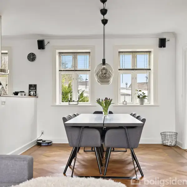 Spisebordet står midt i rummet med seks stole omkring. En vase med tulipaner står på bordet. Bagved ses vinduer, og til venstre er der et køkken.