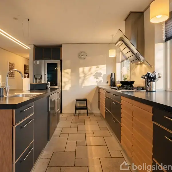 Køkken med to parallelle bordplader; en med håndvask, den anden med komfur. Sollys strømmer ind gennem et vindue. Lyst flisegulv og moderne skabsdesign. Vægur viser kl. 10:10.