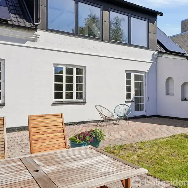 Terrasse med træbord og stole foran hvidt hus. Ved siden af findes to stole ved en dør. Plantekrukker står til højre, omgivet af murværk og græsplæne under blå himmel.