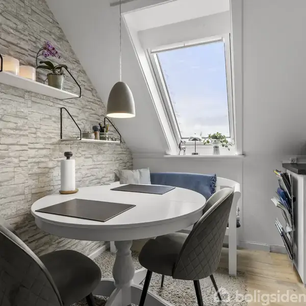 Et rundt, hvidt bord står i et lyst rum med skråt vindue. To stole er placeret omkring bordet, og hylder med dekorative genstande hænger på en stenvæg.