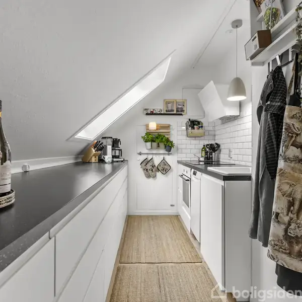 Et langt og smalt køkken med sort bordplade og hvide skabe. Der hænger forklæder og planter på højre væg. Skånke og krydderier står på bordet under et ovenlysvindue.