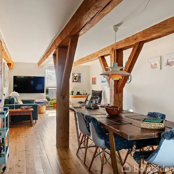 Et langt spisebord er omkranset af stole i en stue med træbjælker. Der er kunst på væggene, en sofa ved et fjernsyn, og bøger på en reol.