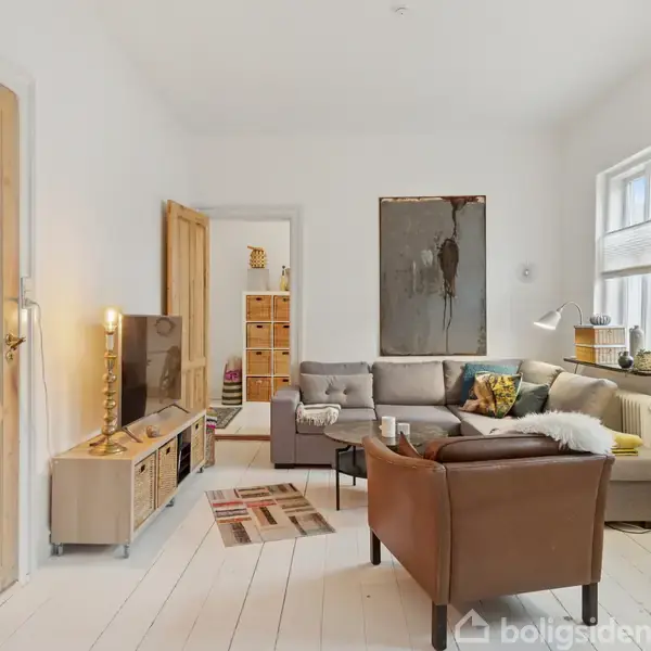 En stue med en grå hjørnesofa og lænestol, omkranset af hvide vægge og trægulv. Et TV står på et skab, bøger på en reol, og en stor plante ved siden af vinduet. Der er kunst på væggen og et tæppe på gulvet.
