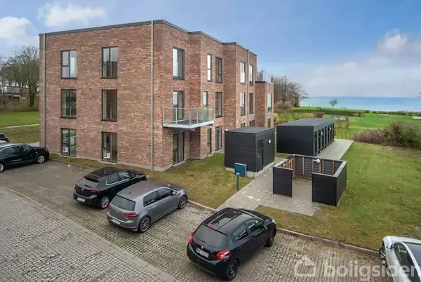 Et moderne, tre-etagers murstensbygning står stille omgivet af parkerede biler og grønne områder. Havet kan ses i baggrunden, hvilket giver et roligt, kystnært miljø.