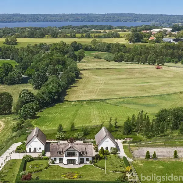Et stort landsted med stråtækte tage står omgivet af velholdte haver. Bagved strækker sig åbne marker og skovklædte områder. I baggrunden ses en stor sø og flere træer.