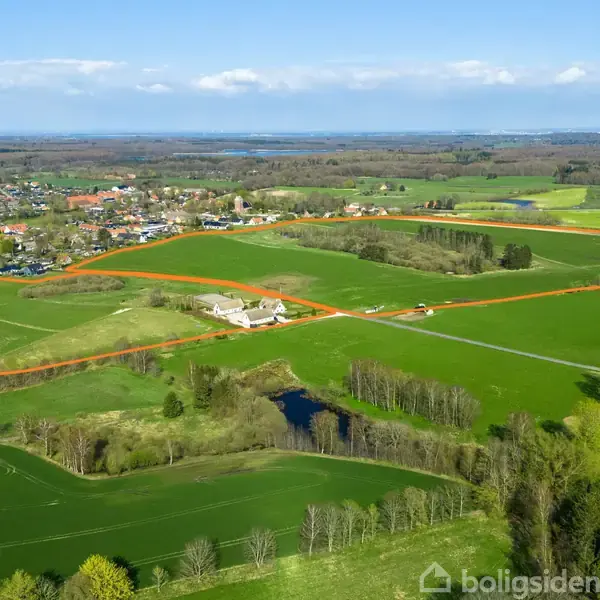 Gårdanlæg, markeret med orange grænse, ligger i et landskabeligt område med grønne marker og små skovområder. En landsby ses i baggrunden under en blå himmel med spredte skyer.