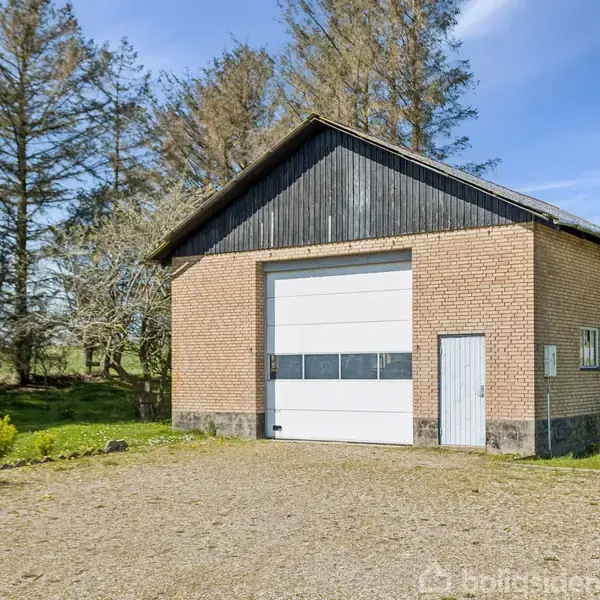 En stor garagebygning af mursten står i et åbent område med græs og træer. Den har en stor port og en lille dør ved siden af.