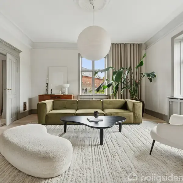 En moderne stue med en grøn sofa, hvor et rundt, hvidt loftlys hænger. Rummet indeholder også en hvid lænestol, et ovalt, sort sofabord og store vinduer med udsigt til en bygning.