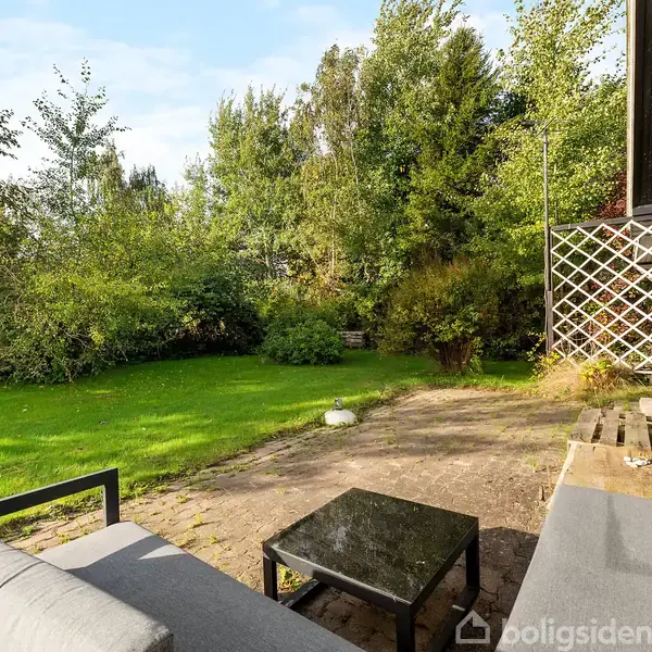 En grå sofa og et sort bord står på en stenbelagt terrasse ved siden af et sort træhus, omgivet af en grøn have med høje træer og buske.