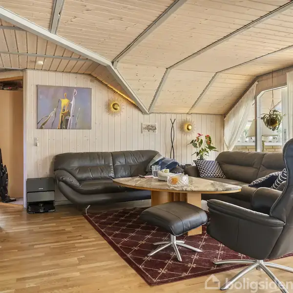 Stue med sorte lædersofaer og et rundt sofabord i træ, omgivet af store vinduer og skråt loft i træ. På væggen hænger et billede. Blomster pynter rummet.