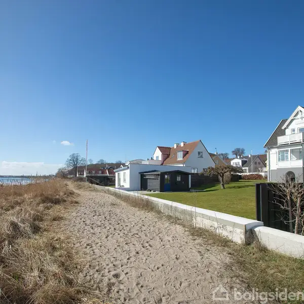 Strandvej med grussti løber langs vandet. Huse med haver ligger på højre side, omgivet af klar blå himmel.