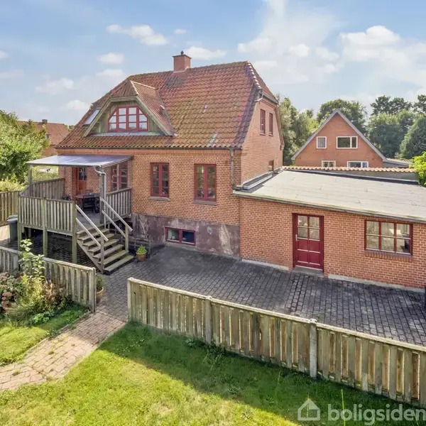En murstenshus med rødt tag ligger i en have med træhegn og flisebelagt gårdsplads. Trætrappe fører op til verandaen. Blå himmel og grønne buske i baggrunden.