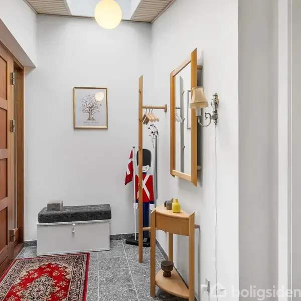 En entré med gråt flisegulv og en rød løber, hvor en dør er til venstre, et spejl hænger til højre, og et skab og flag står i hjørnet.