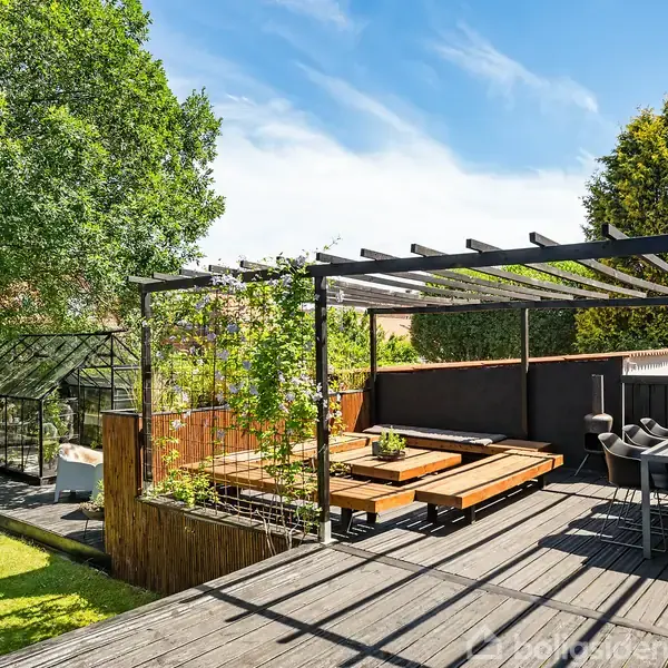 Terrasse med pergola og spisebord, beliggende i en grøn have med græsplæne. Et drivhus er synligt i baggrunden. Solrigt vejr og blå himmel skaber en afslappende atmosfære.