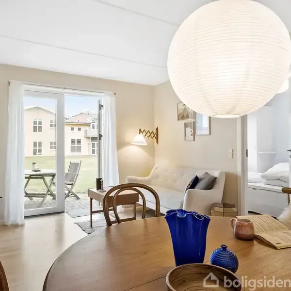 Rundt spisebord med blå vase står i en lys stue. Sofa med puder ses ved siden af store glasdøre, der fører til en balkon. Store papirlamper hænger fra loftet.