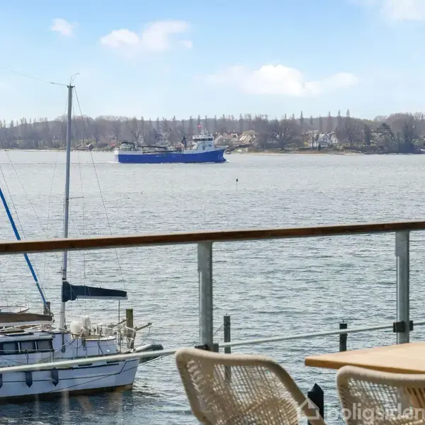 Et blåt skib sejler på en bred flod med skovklædte bredder i baggrunden. I forgrunden ses en lystbådehavn med både fortøjet ved en anløbsbro.