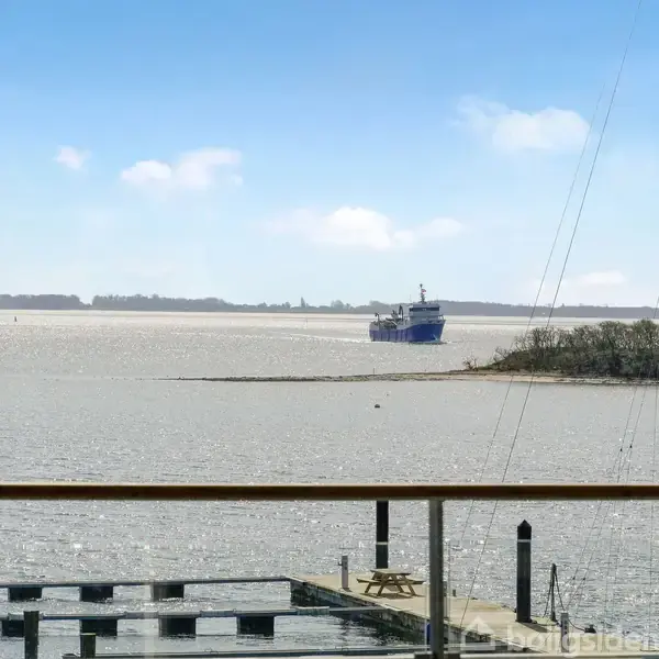 Skib sejler på bred flod, set fra glasvindue. Træbro i forgrunden, rolig vandflade og kystlinje i baggrunden under en blå himmel.