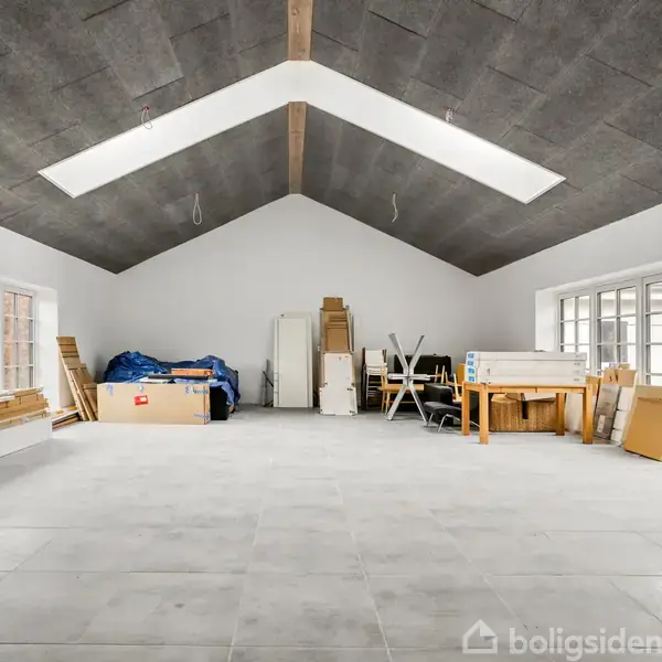 Et rummeligt, lyst rum med højt til loftet indeholder opstablede byggematerialer og møbler. Store vinduer langs væggene tillader dagslys at strømme ind, hvilket skaber en åben atmosfære.