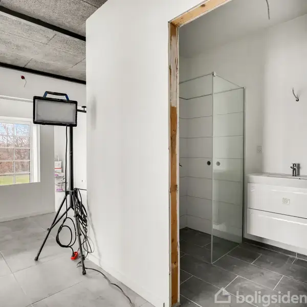 Et badeværelse med glasbruser og hvid vask, set gennem en åben dør. En stue med store vinduer og en oplyst projektør på stativ står ved siden af døren.