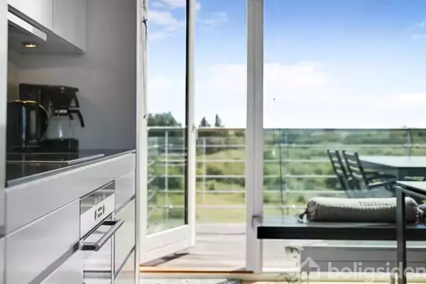 Et køkken med en kaffemaskine står ved siden af åbne altandøre, der fører ud til en altan med udsigt over frodigt, grønt landskab.