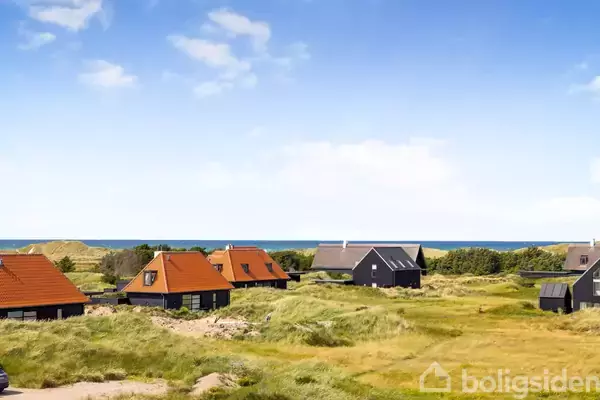 Huse med røde og sorte tage står spredt på en græsmark. Havet og klitterne ses i baggrunden under en blå himmel med få skyer.