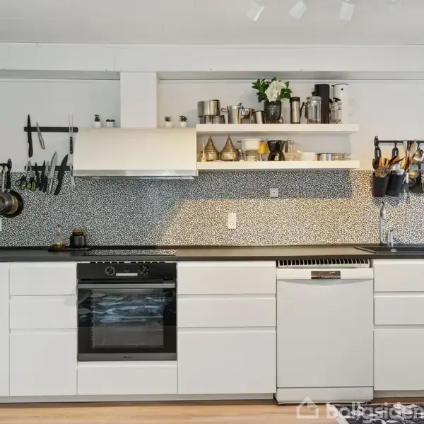 Et spisebord med fem sorte stole står i et lyst rum. Bordet har en vase med blomster. Væggene har billeder, og et vindue giver udsigt til en murstensvæg udenfor.