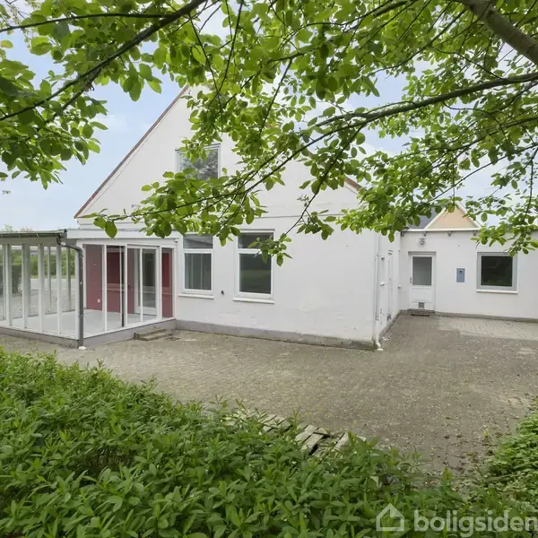 Hus med hvid facade og store vinduer, står på en flisebelagt gård. Omkranset af grønne buske og træer.