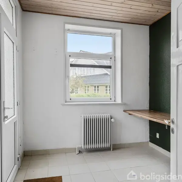 En lys entré med en radiator under vinduet, et lille træbord til højre og en glasdør til venstre. Udsigt til et hus og have udenfor.