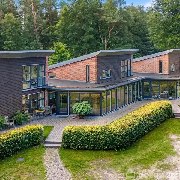 Et moderne huskompleks med flade tage og store vinduespartier er placeret i en skovklædt område. Bygningerne omgives af velplejede buske og en stenbelagt sti foran dem.