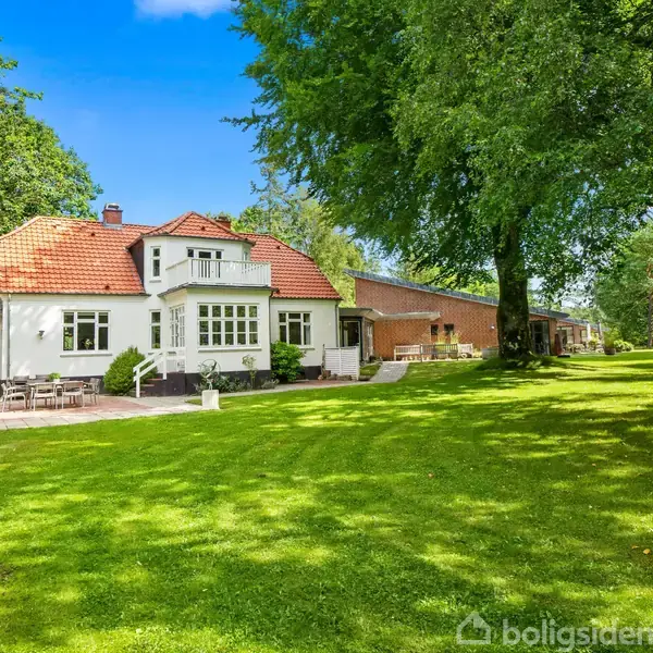 Et hvidt hus med rødt tag står i en grøn have. I haven er der store træer, og en udendørsterrasse ses til venstre for huset.