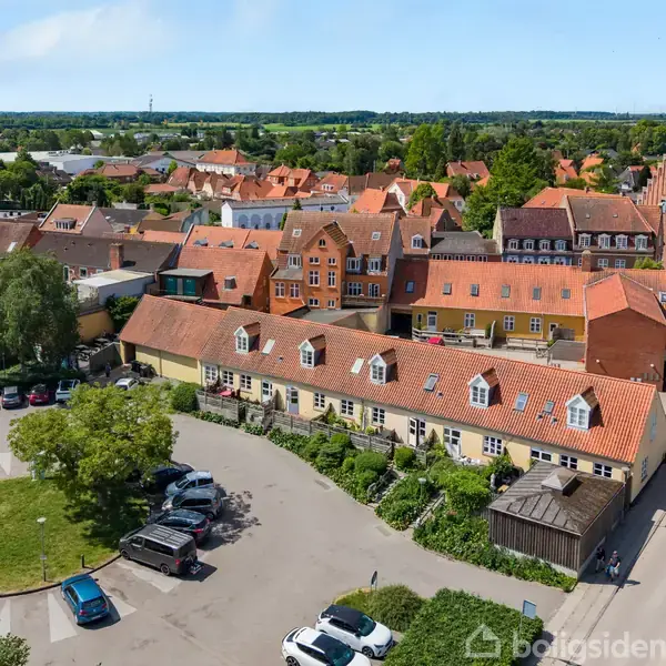 Flere huse med røde tage står tæt sammen; en parkeringsplads med biler ses foran. I baggrunden er der flere bygninger og grønne træer under en klar himmel.