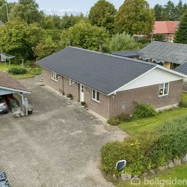 Et murstenshus med mørkt tag ligger ved en brostensbelagt indkørsel. En carport til venstre indeholder en parkeret bil. Haven er omkranset af grønne træer og buske.