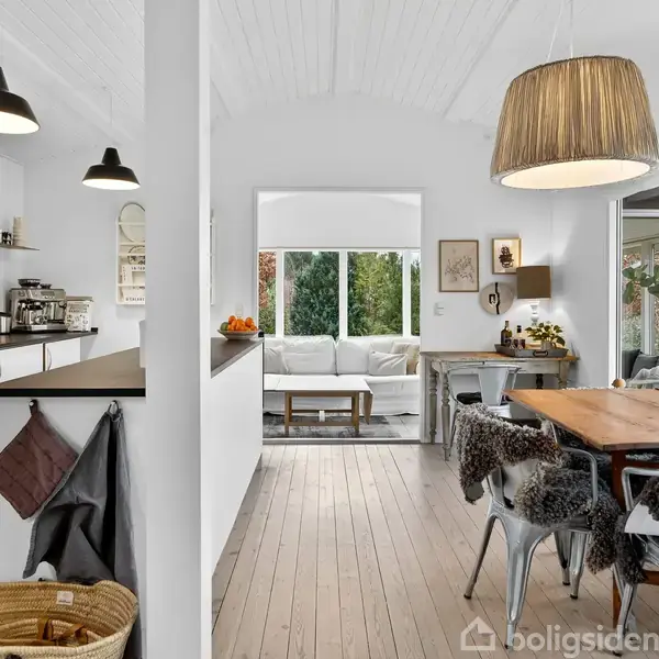 Et moderne køkken-alrum er indrettet med hvidmalede vægge og trægulv. Et spisebord med stole i forgrunden, køkkenborde langs væggen til venstre og en sofa i den bageste del.