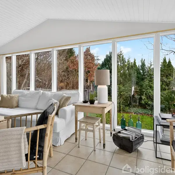 Lys stue med hvid sofa og sofaborde, omgivet af store vinduer med udsigt til frodig have. En gulvlampe og planter står ved siden af sofaen.