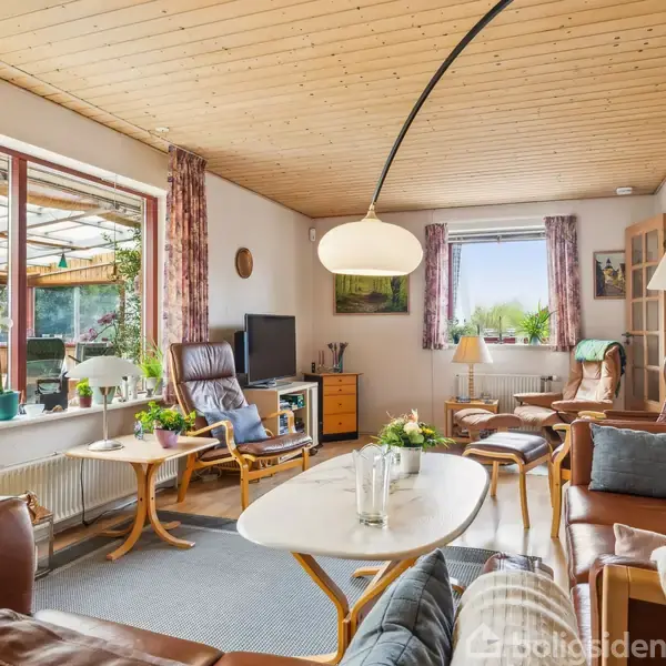 En stue med brune lædersofaer står omkring et hvidt ovalt sofabord. Rum oplyst af stor vinduesparti med planter i vindueskarmen, træloft og tæppe på gulvet.