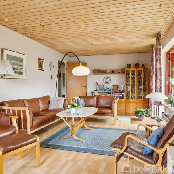 Brune lædermøbler står i en stue med trægulv og træloft, omkranset af store gardindækkede vinduer. En reol med glasdøre indeholder bøger og pynt. En buet lampe lyser op.