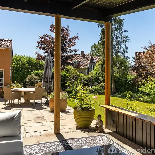 Terrasseområde med sofa under overdækning. Træbord og stole står udenfor nær en gul bygning. Omgivet af frodig have med græsplæne og træer under blå himmel.