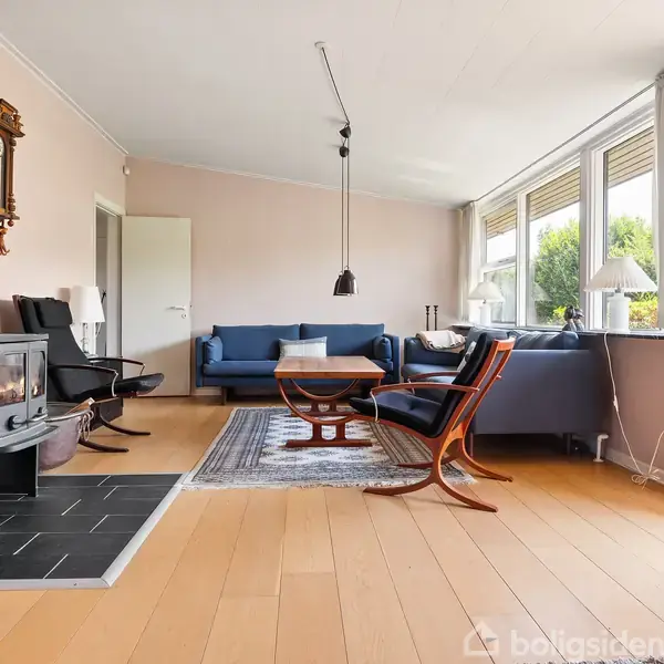 Balkon med flisebelagt gulv åbner op mod en murstensbygget væg med en glasdør. I baggrunden ses grønne træer under en blå himmel med skyer.