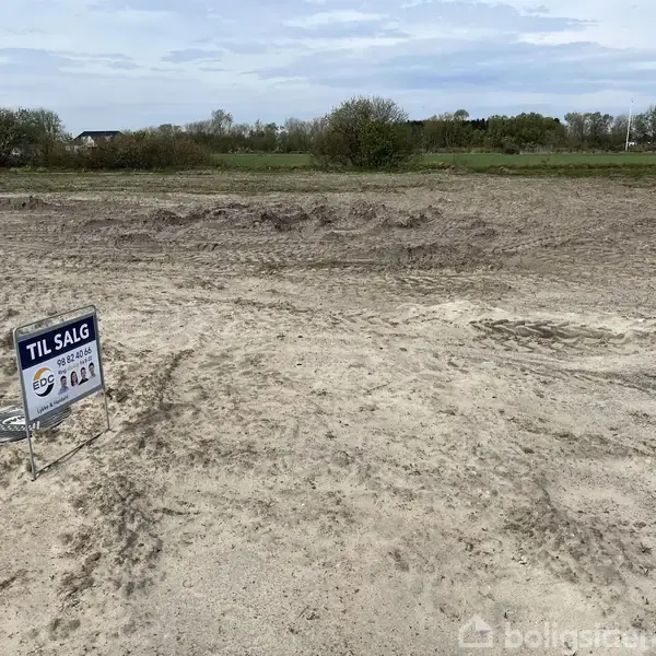 Skilt står på bar jordgrund, der er til salg, omgivet af græsmark og træer. Tekst på skilt: "TIL SALG, 3G Bolig, Jonas Max.”