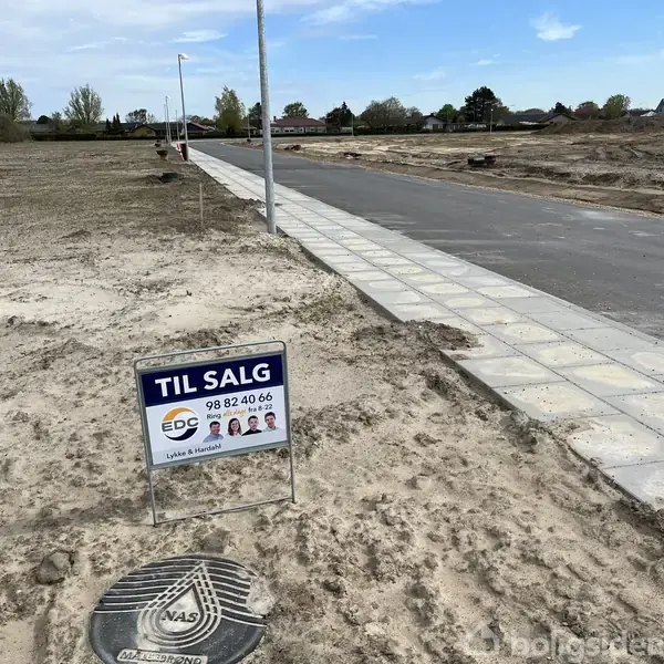 Et skilt med teksten "TIL SALG" og telefonnummeret 98 82 40 66 står ved siden af en nyanlagt vej i et åbent, ubebygget område.