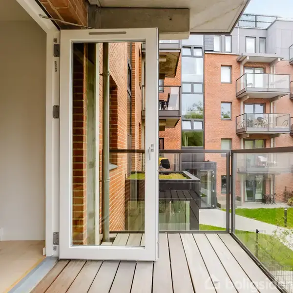 En altandør står åben og fører ud til en balkon med glasrækværk. I baggrunden ses en moderne murstensbygning med flere altaner samt en græsplæne og gangsti.