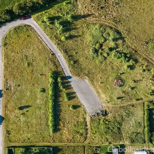 En landevej danner en kurve mellem åbne græsarealer med områder af buske og træer. Til venstre ses en række moderne bygninger og haver langs vejen under en klar blå himmel.
