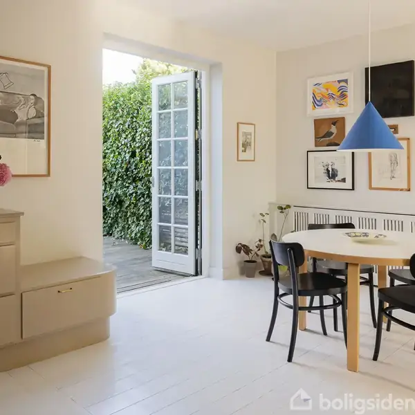 Rundt spisebord med fire sorte stole står i et lyst rum. Væggene har kunstneriske billeder, en blå lampe hænger over bordet. En dør åbner til en terrasse med grøn beplantning.
