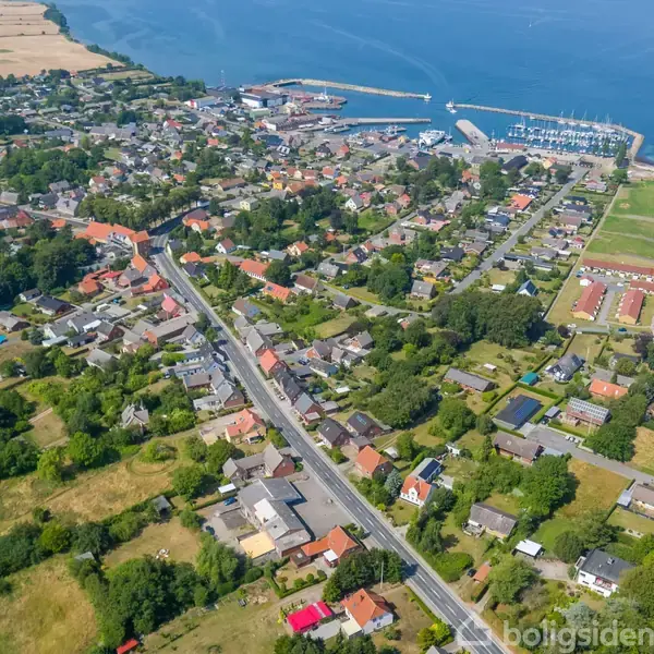 En by med huse langs en vej, som fører til en havn ved stranden. En blå pil peger på et bestemt hus omgivet af træer og marker nær kysten.