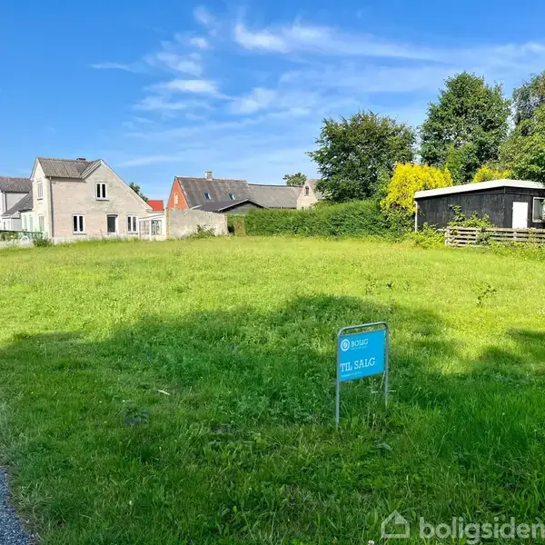 Græsareal til salg, beliggende ved siden af stier og boliger. Et skilt med teksten "EDC Til Salg" står centralt. Omkring er der træer og en lille bygning.