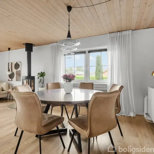 Et rundt spisebord med seks beige stole står i en lys stue. Ved bagvæggen er vinduer med hvide gardiner, og rummet har også en sofa og en brændeovn.