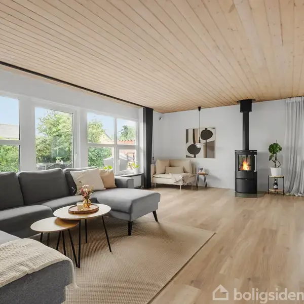 Stue med grå hjørnesofa, små runde borde med dekorationer, træbrændeovn tændt i hjørnet, store vinduer giver naturligt lys. Trægulv og træloft, minimalistisk kunst hænger på væggen.