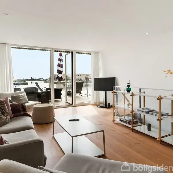 Stue med sofaer står omkring et sofabord; glasskabet til højre indeholder dekorationer. Store vinduer viser en balkon med borde og stole. Et TV står mod væggen ved balkonen.
