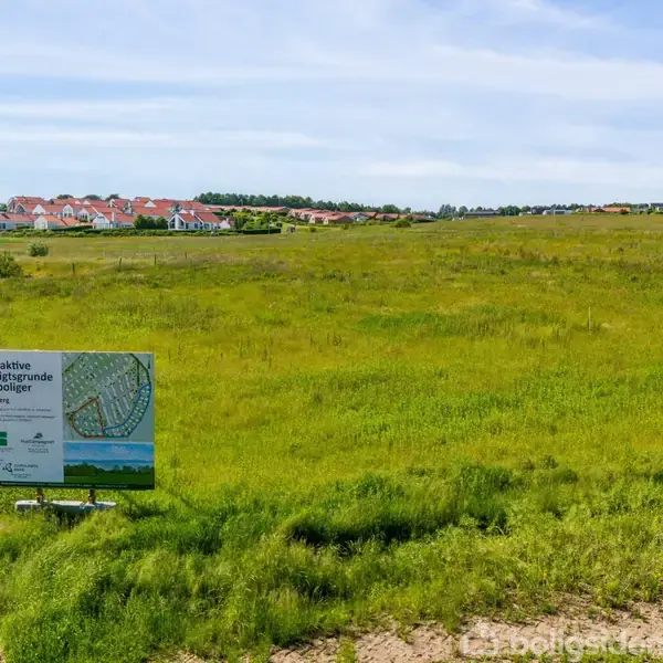 En reklameskilt står på en grøn mark; teksten lyder: "Attraktive udstykninger og boliger, Stilling." I baggrunden ses et boligområde med rødt tag.