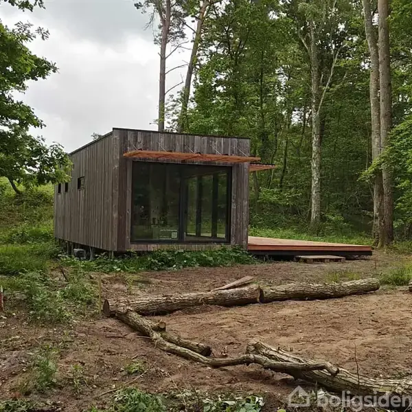 En lille moderne træhytte står i en skov, med store glasvinduer og en træterrasse. Omgivet af træer og grene, skaber det en afsondret og naturlig atmosfære.